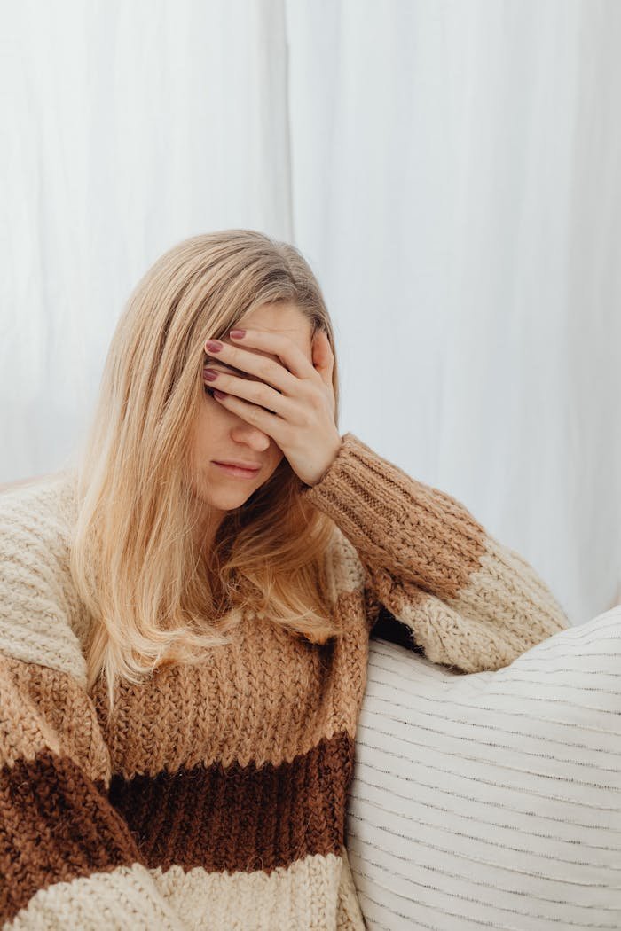A woman in a warm sweater covers her face, appearing unwell while sitting indoors.
