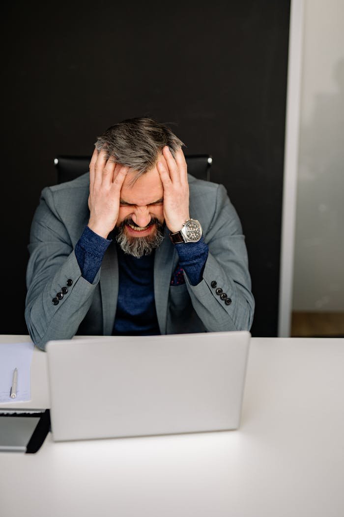 Stressed businessman in gray suit with head in hands at office desk, facing laptop.