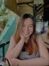Portrait of a smiling woman in a sleeveless shirt, seated at a caf&eacute; with a tropical backdrop.