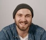 Close-up portrait of a cheerful young man with a beard and knit cap wearing denim.